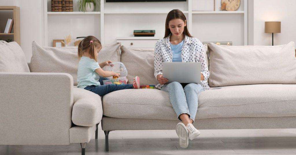 A young mom sitting on a couch and working on her laptop. Her young daughter is playing with toys beside her.