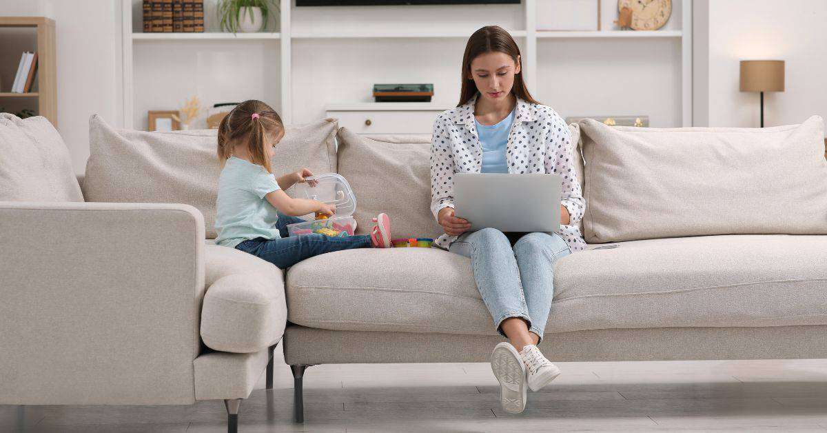 A young mom sitting on a couch and working on her laptop. Her young daughter is playing with toys beside her.